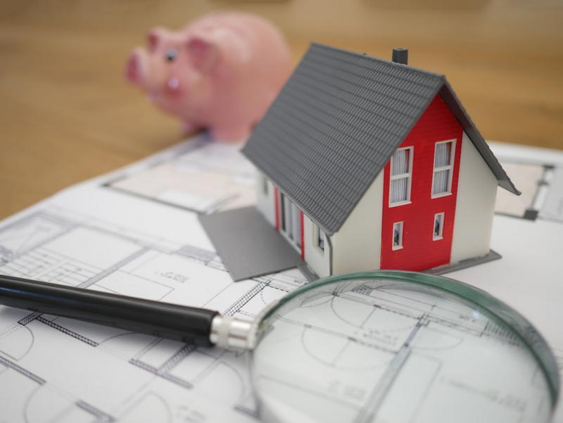 A model house, magnifying glass, pen, and piggy bank are placed on an architectural blueprint.